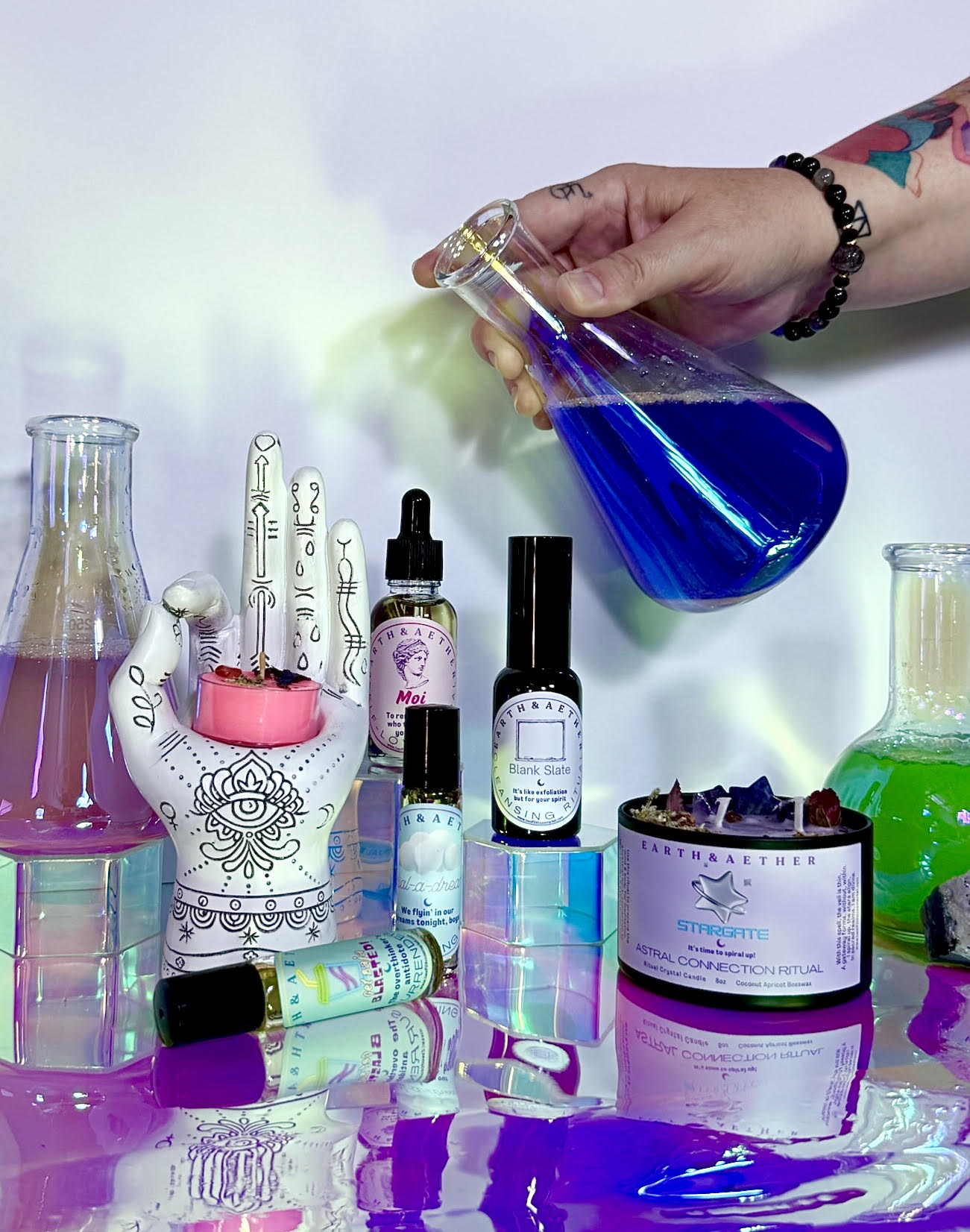 Earth & Aether oils, sprays and a Stargate ritual candle beside a palmistry hand, with a flask of blue potion being poured